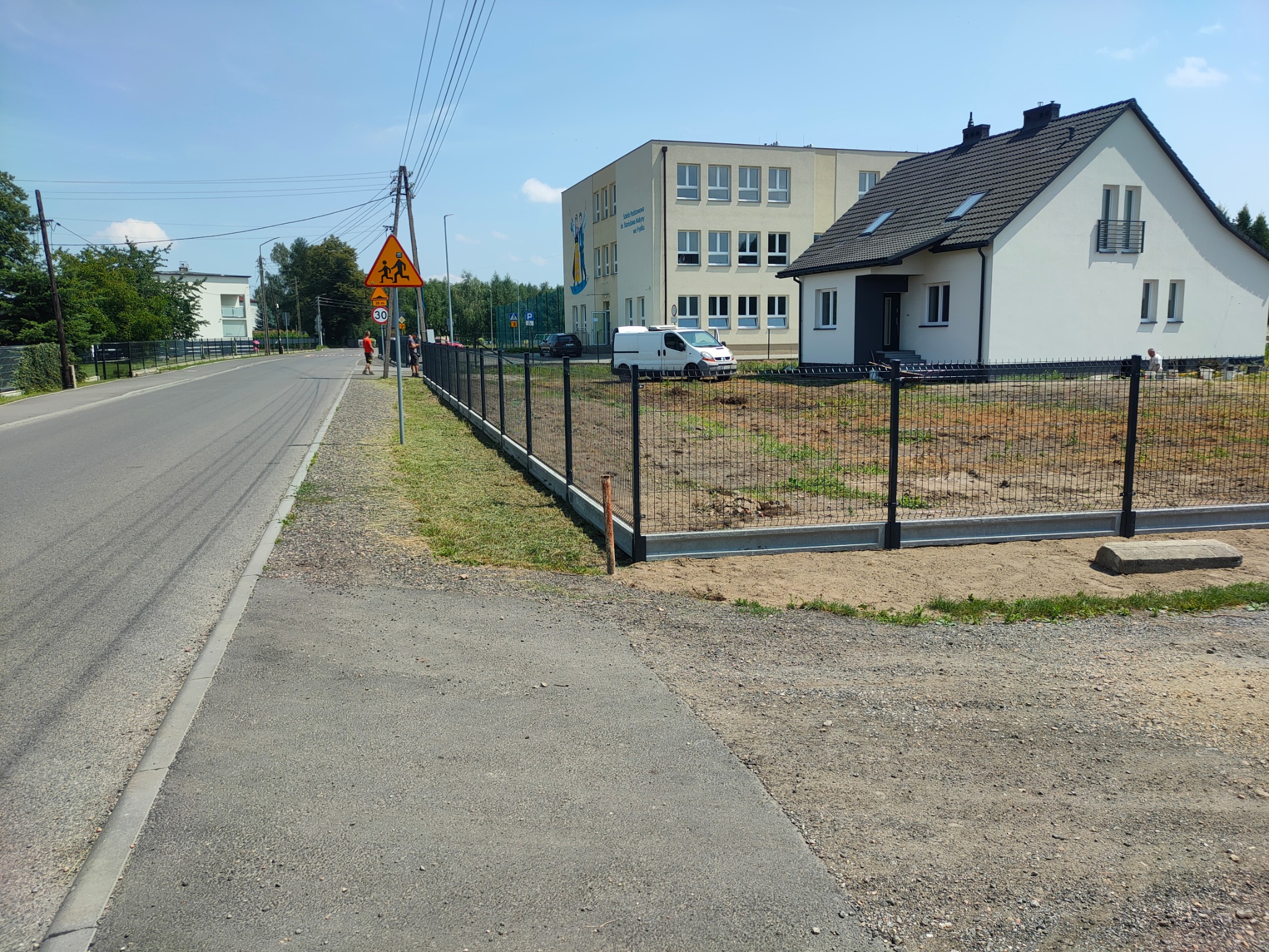Nowo postawione, czarne ogrodzenie panelowe z betonową podmurówką oddziela posesję z domem i budynkiem szkoły od ulicy z chodnikiem i znakiem ostrzegawczym.