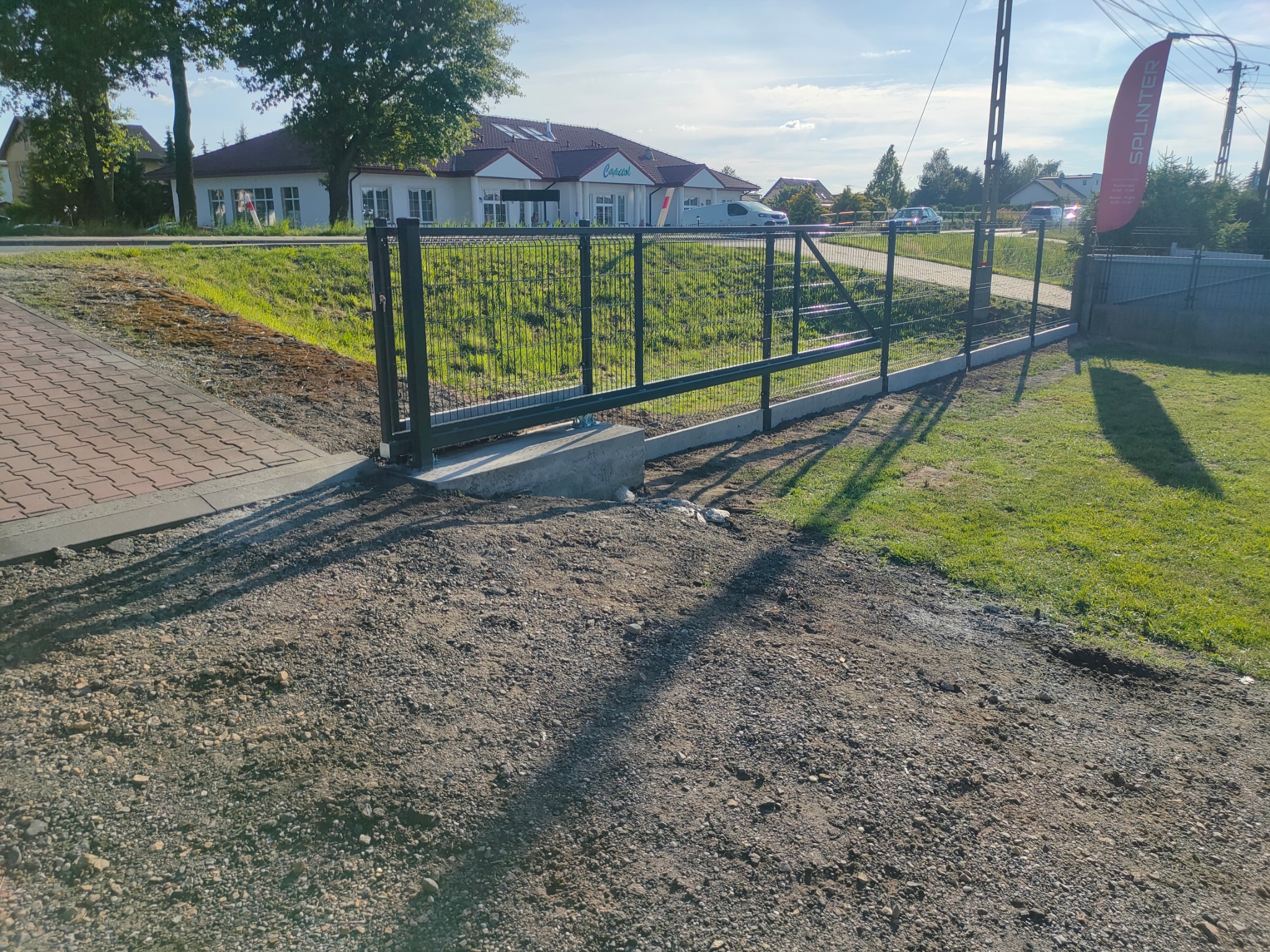 Nowo zainstalowana, nowoczesna, metalowa brama wjazdowa z siatkowym wypełnieniem, osadzona na betonowych fundamentach, otoczenie w trakcie wykończenia, widoczna kostka brukowa i budynek w tle.
