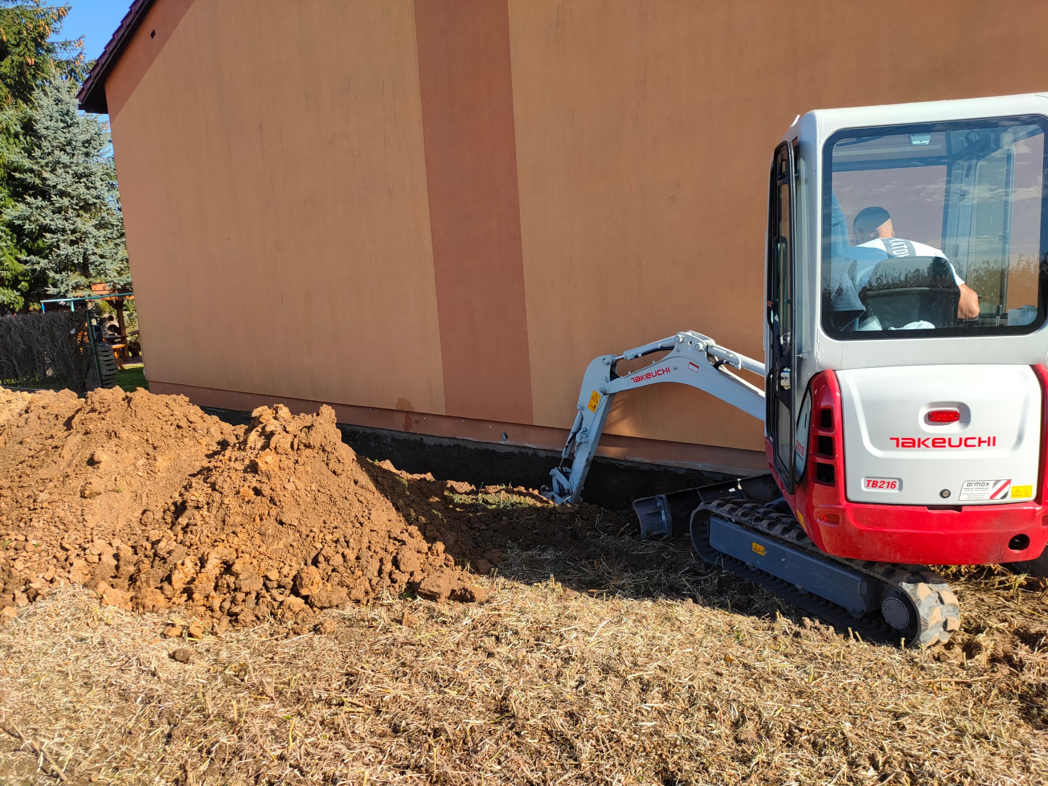 Mini koparka Takeuchi TB216 wykonuje wykop w ziemi przy ścianie budynku w Porębie, obok leży sterta wykopanej ziemi.