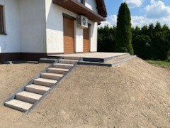 Ułożone schody z kostki brukowej prowadzące do tarasu przed domem, teren wokół częściowo wyrównany, widoczny budynek z brązową bramą garażową i klimatyzatorem.