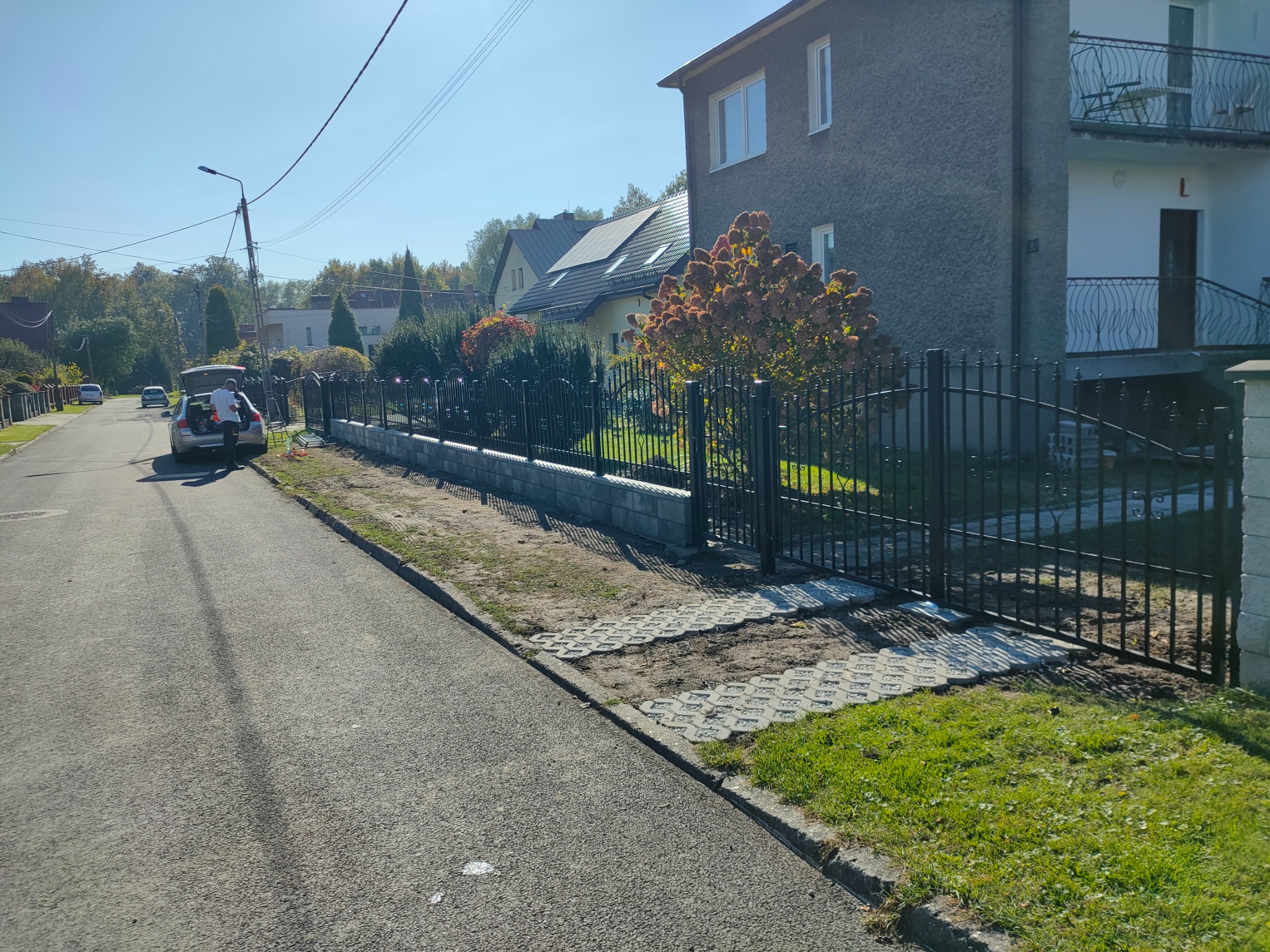 Nowoczesne, czarne ogrodzenie panelowe z bramą przesuwną, zamontowane na betonowej podmurówce, widoczne od strony ulicy, z samochodem i osobą przy nim. Na posesji widoczny dom z balkonem i krzewy.