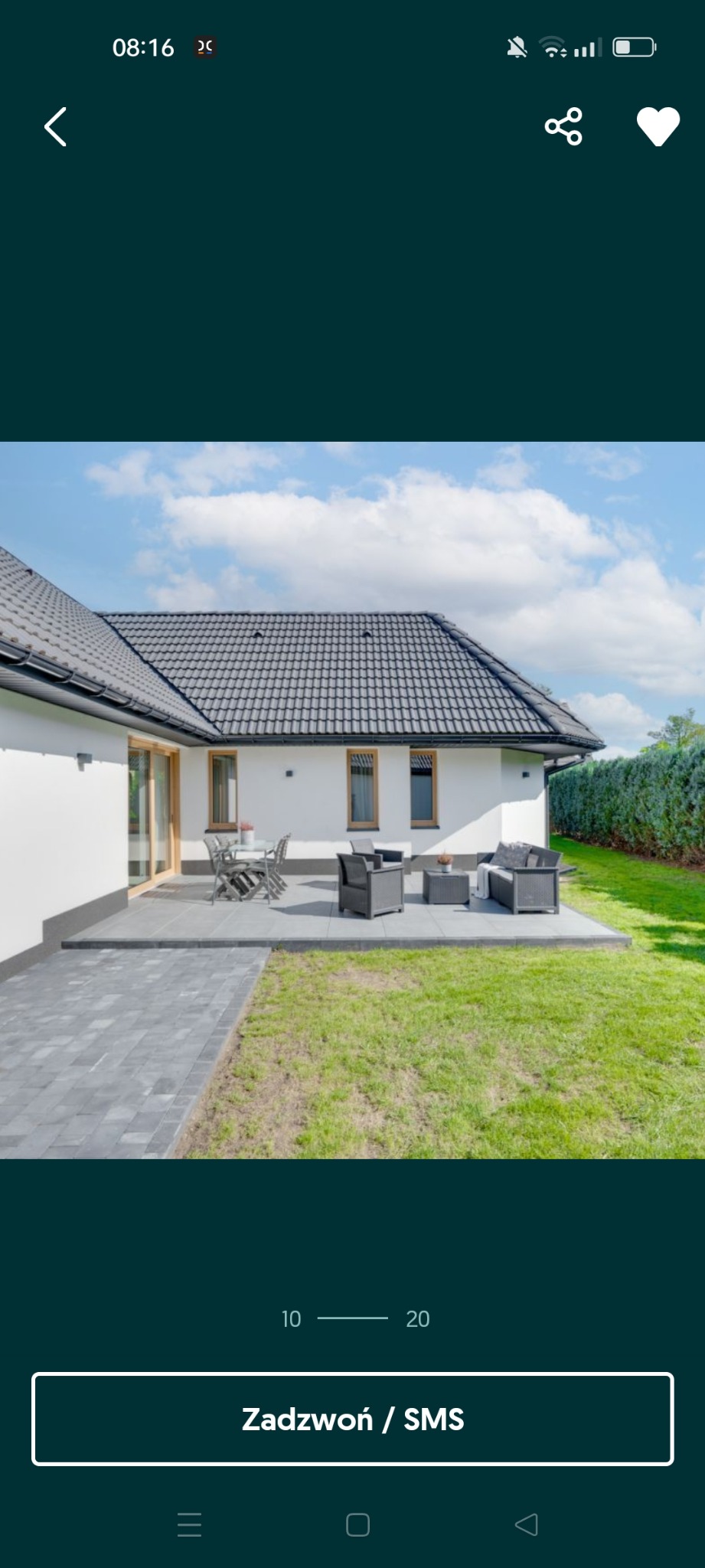 Nowoczesny taras z szarej kostki brukowej przy białym domu z ciemną dachówką, otoczony zielonym trawnikiem i tujami.