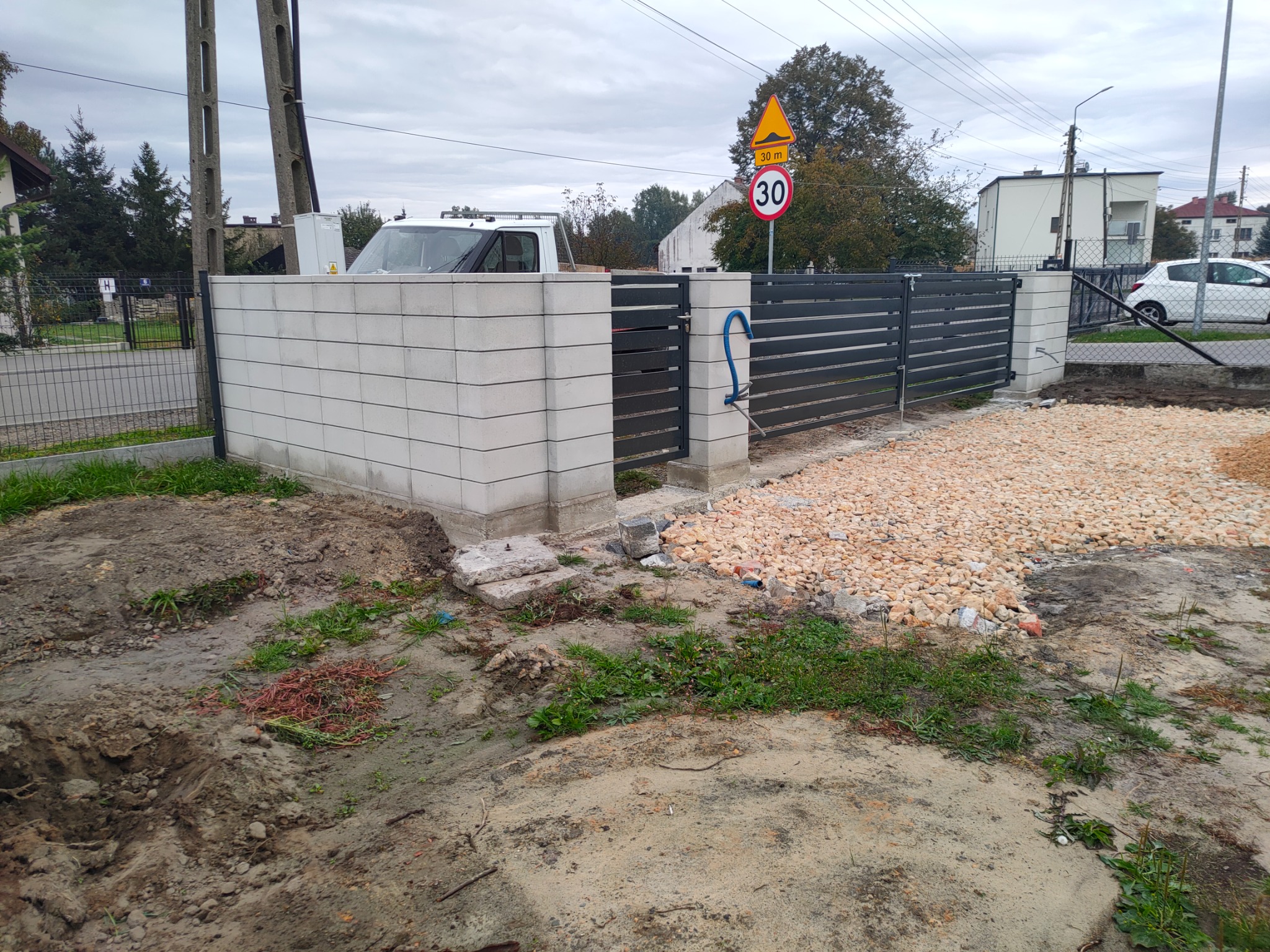 Nowoczesne ogrodzenie panelowe z betonowych bloczków, częściowo ukończone, z widocznym gruzem i ziemią, znak drogowy ograniczenia prędkości w tle.