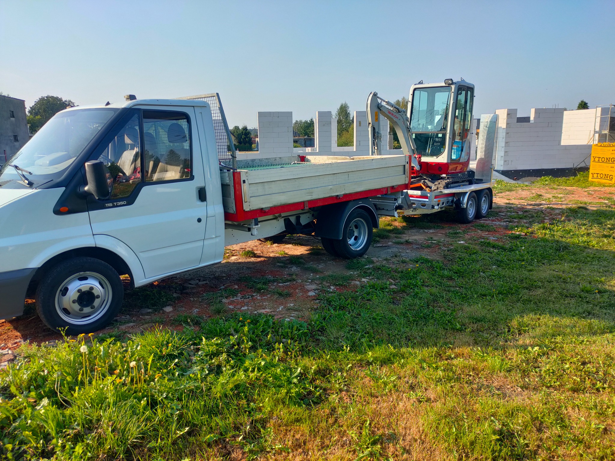 Biały samochód dostawczy Ford Transit T350 z przyczepą przewożącą mini-koparkę Takeuchi na tle częściowo wzniesionego budynku z białych bloczków Ytong.