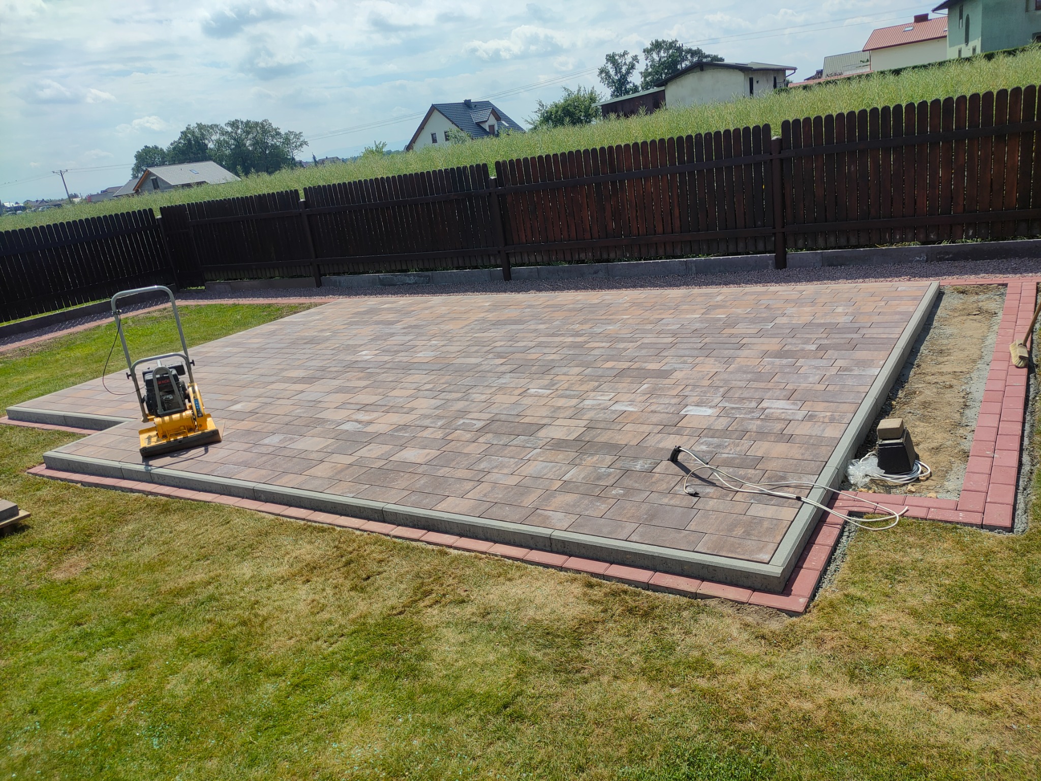 Ułożona kostka brukowa w odcieniach brązu i szarości, obrzeża z czerwonej kostki, ubijarka wibracyjna na powierzchni, widok z góry na trawnik i ogrodzenie.