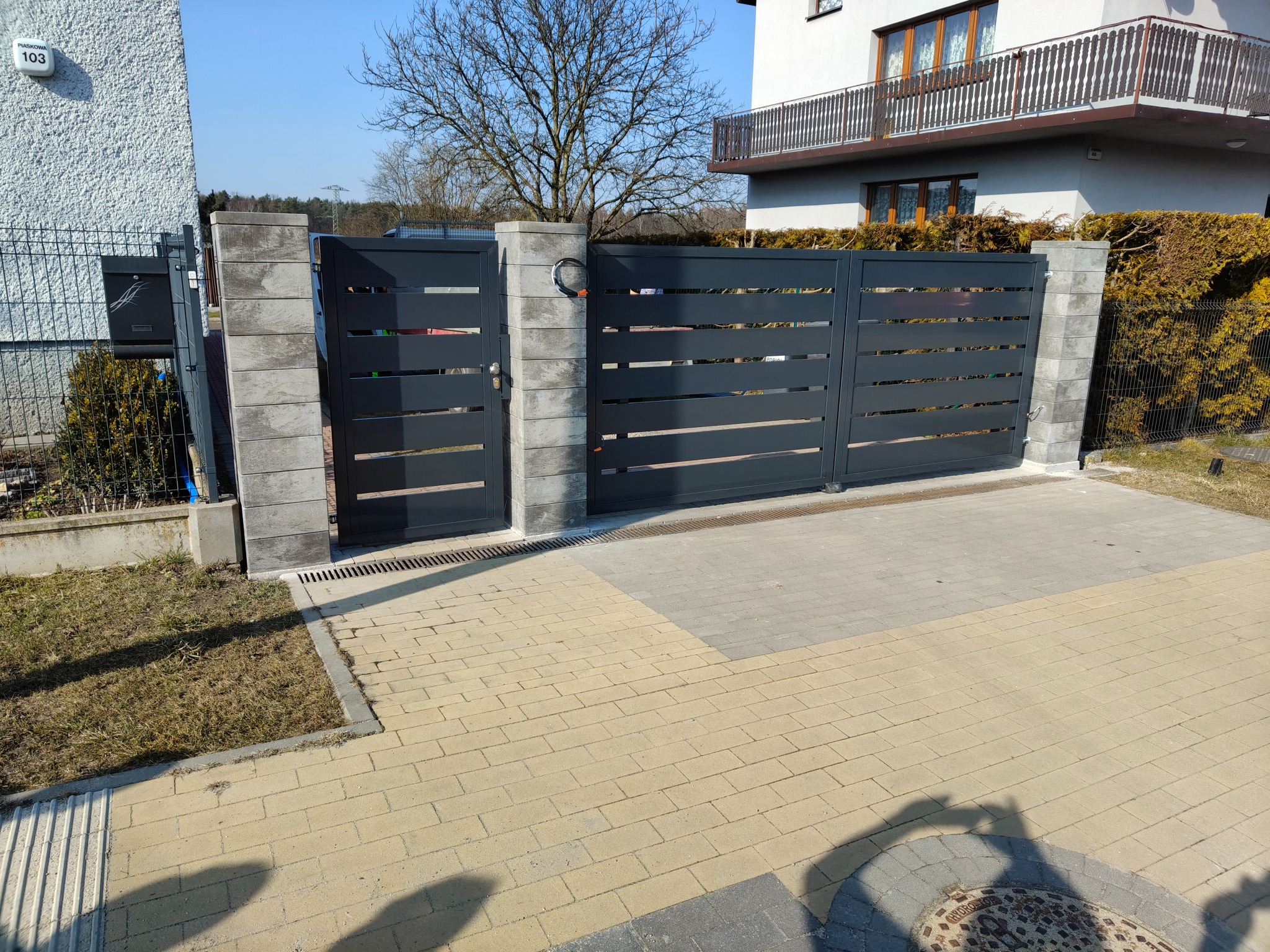 Nowoczesna, pozioma brama wjazdowa z szarego metalu, osadzona na słupkach z szarej kostki, na tle domu z balkonem i kostki brukowej w dwóch odcieniach.