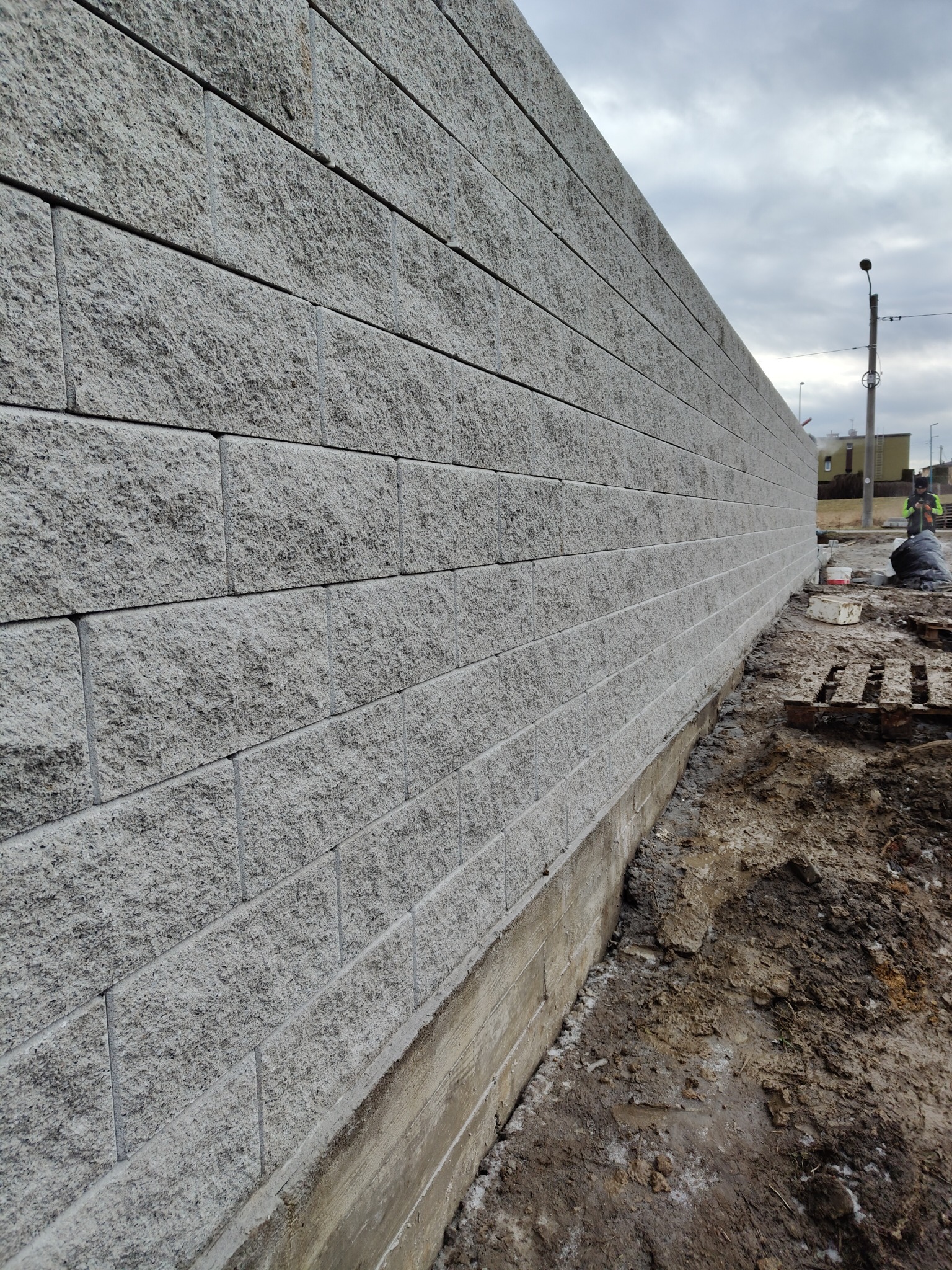 Szara ściana oporowa z betonowych bloczków, widoczna z boku, z fundamentem i ziemią u podstawy. W tle niebo i budynki.