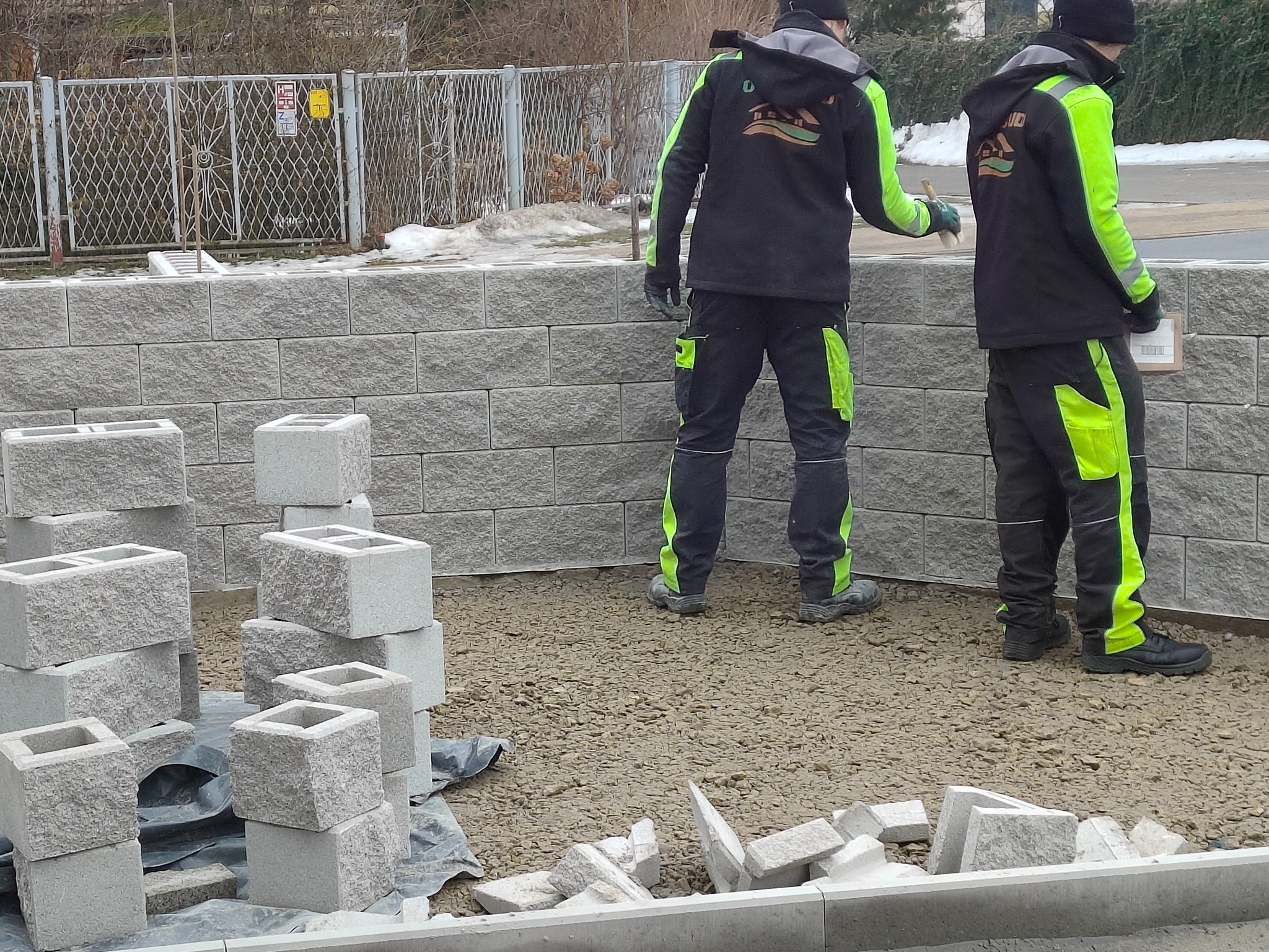 Budowa muru z szarej kostki betonowej. Dwóch pracowników w odzieży roboczej z neonowymi wstawkami. Częściowo ukończony mur i przygotowane elementy do dalszej budowy.