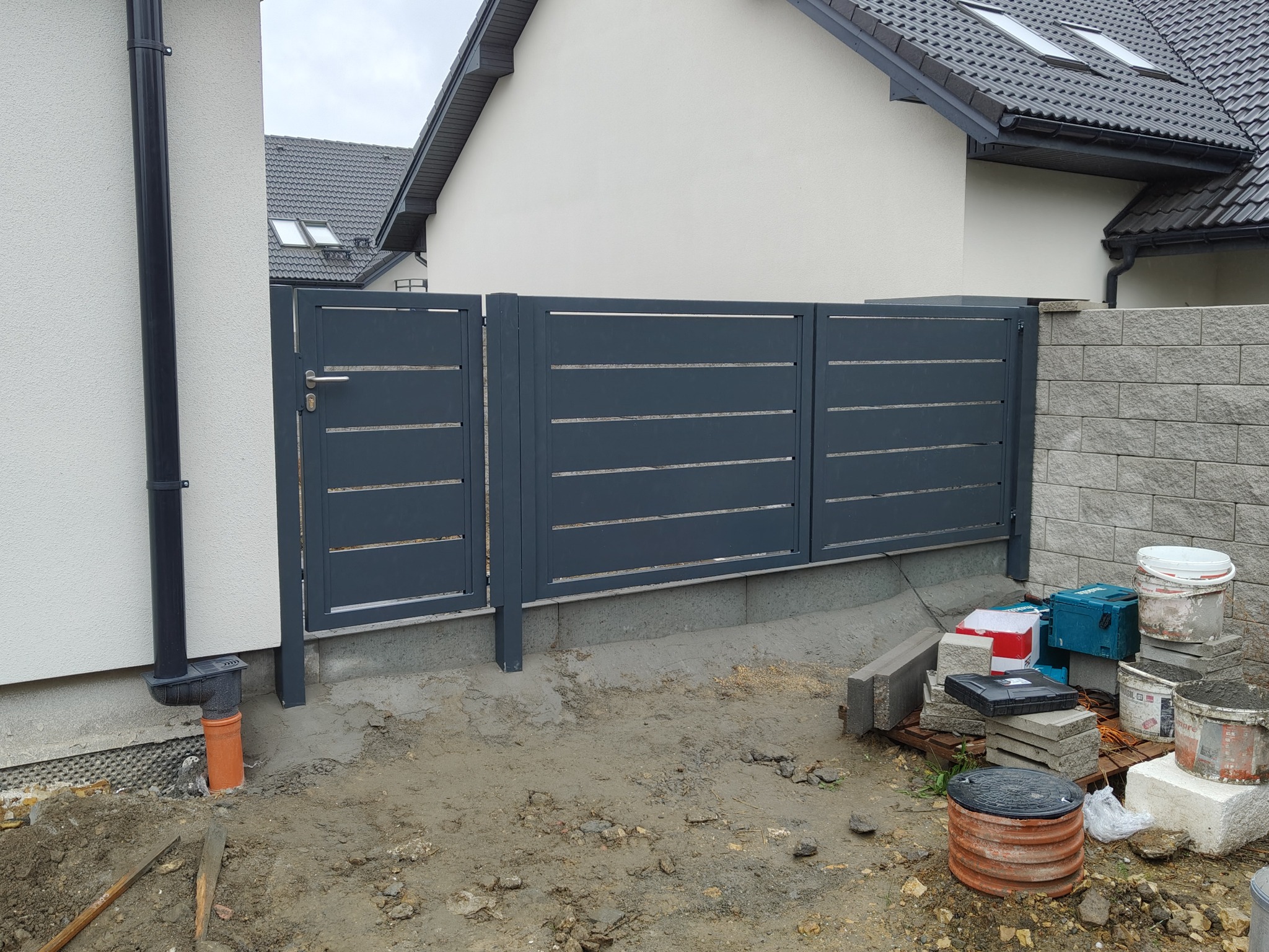 Nowoczesne, poziome przęsła ogrodzeniowe z szarego metalu, zamontowane na betonowej podmurówce. Widoczne narzędzia i materiały budowlane. Tło stanowi ściana budynku.