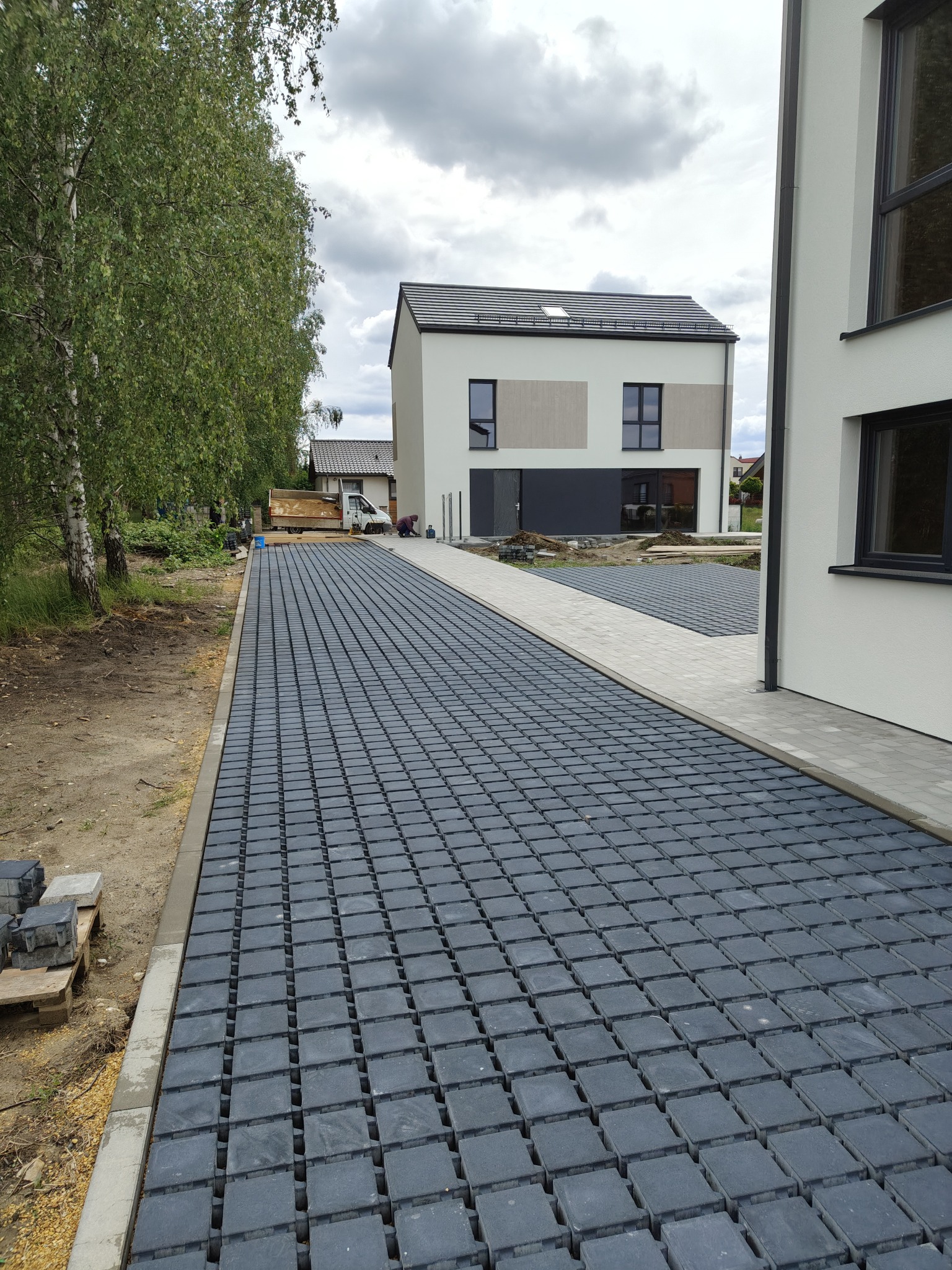 Ułożona, czarna ażurowa kostka brukowa na podjeździe do nowoczesnego domu. Widoczne palety z kostką i prace wykończeniowe. Pochmurne niebo.