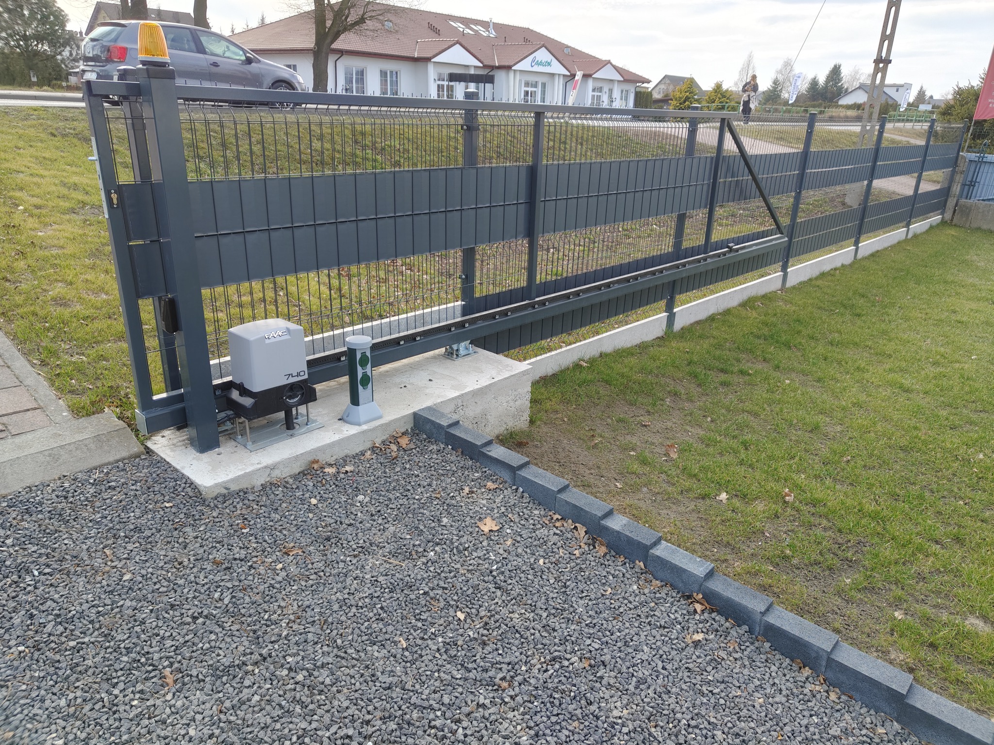 Nowoczesna, przesuwna brama w kolorze grafitowym z automatyką, zamontowana na posesji z trawnikiem i szarą nawierzchnią z kamyków. Widoczny napęd bramy.