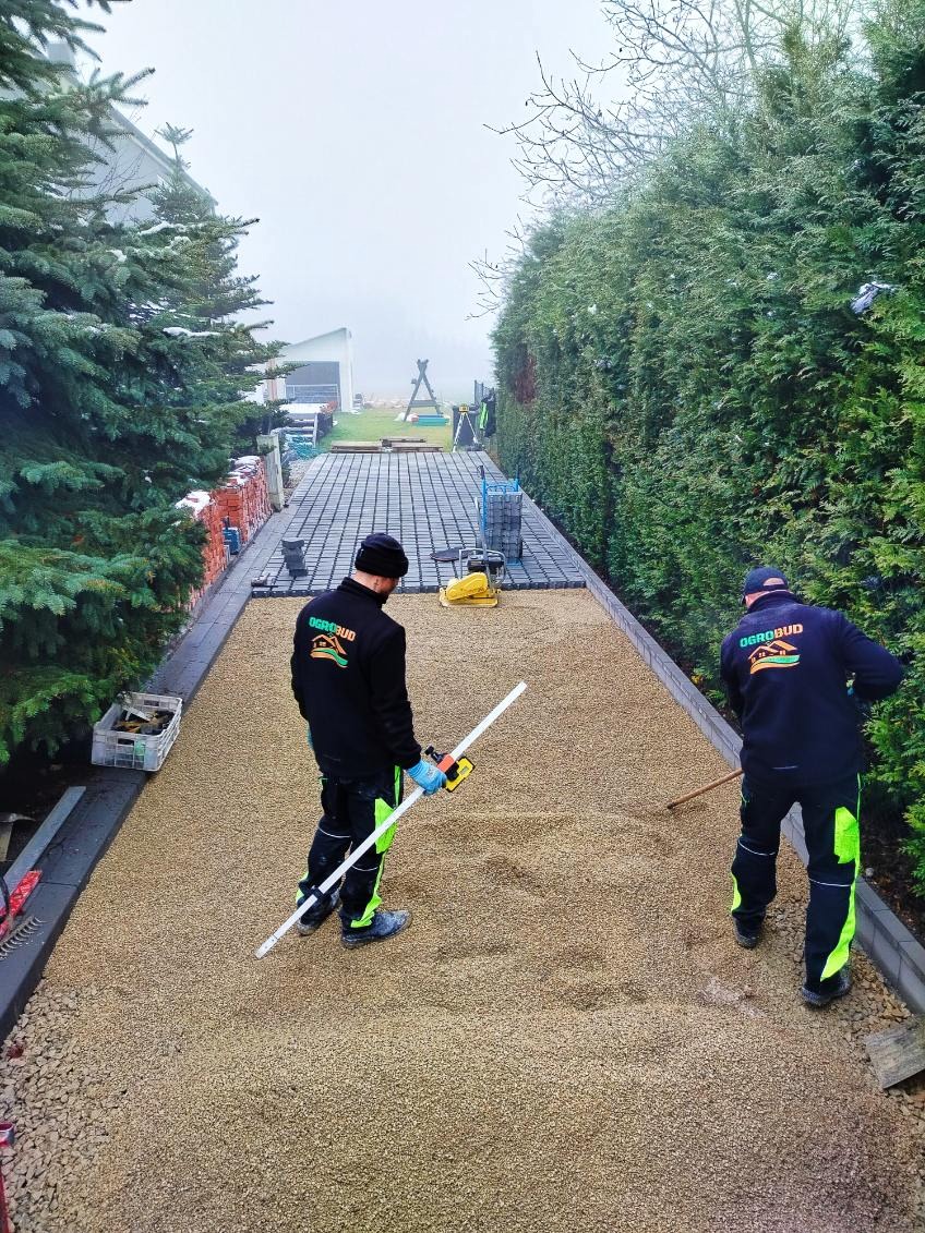 Pracownicy w trakcie układania podbudowy pod kostkę brukową. Widoczne narzędzia i materiały budowlane. Tło: zielony żywopłot i drzewa iglaste.