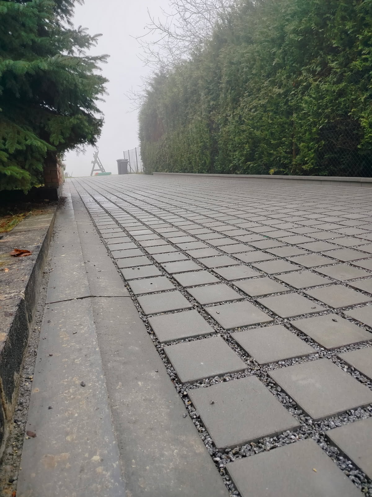 Szara kostka brukowa ułożona w kratę, wypełniona drobnym żwirem, prowadząca w górę w kierunku zielonego żywopłotu i drzew w mglisty dzień. Widok z dołu.
