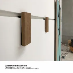 System zawieszania obrazów w galerii, widoczne drewniane uchwyty mocujące obrazy do ściany, w tle częściowo widoczny obraz w jasnych kolorach i fragment podłogi z kablami.