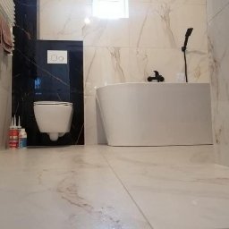 Usługi glazurnicze Binex - Wykończona łazienka z wanną i toaletą w zabudowie, płytki imitujące marmur, czarna armatura, akcesoria montażowe widoczne na podłodze.