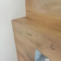 Fragment łazienki: drewnopodobna okładzina ścienna z przyciskiem spłuczki, białe płytki. Minimalistyczny design, ciepłe odcienie drewna, nowoczesne wykończenie.