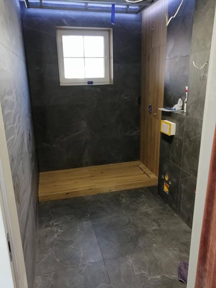 Nowoczesna łazienka w trakcie remontu: ciemne płytki imitujące kamień, drewniany brodzik i białe okno. Widoczne elementy instalacji pod oświetleniem sufitowym.