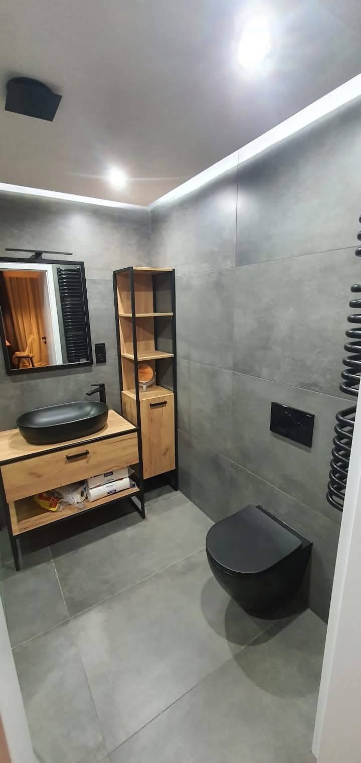 Stylowa łazienka w szarościach z drewnianymi akcentami. Umywalka nablatowa, wisząca toaleta i grzejnik drabinkowy w czarnym kolorze.