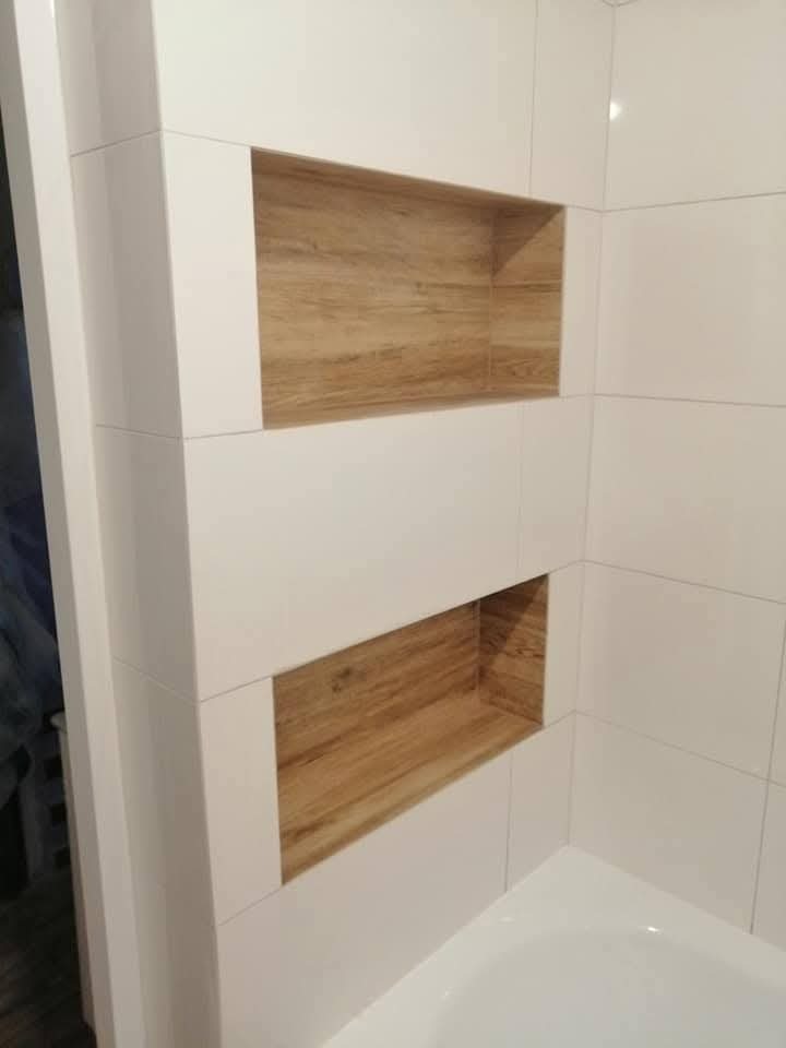 Białe płytki w łazience z dwoma wnękami wyłożonymi drewnopodobnymi płytkami, widok na narożnik wanny i fragment futryny drzwi.