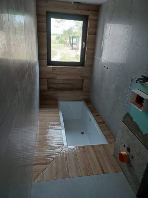 Wnęka z oknem w łazience wyłożonej płytkami imitującymi drewno. Widoczna wanna, fragment instalacji i surowe, szare płytki na ścianach. Nietypowe połączenie materiałów.