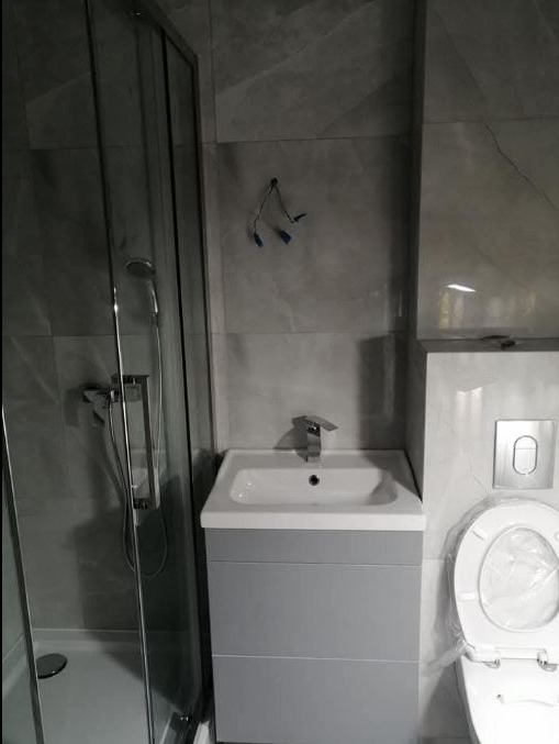 Szara łazienka z kabiną prysznicową, umywalką z szafką i toaletą. Widoczne szare płytki na ścianach. Na ścianie wystają kable.