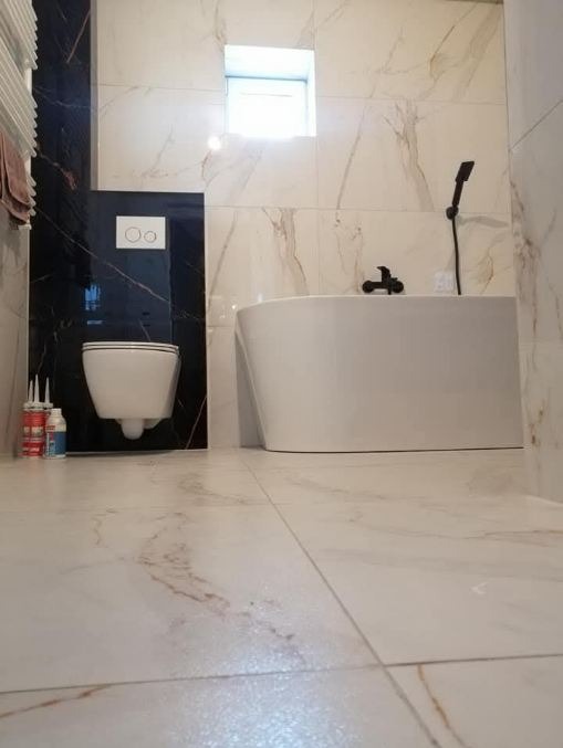 Wykończona łazienka z wanną i toaletą w zabudowie, płytki imitujące marmur, czarna armatura, akcesoria montażowe widoczne na podłodze.