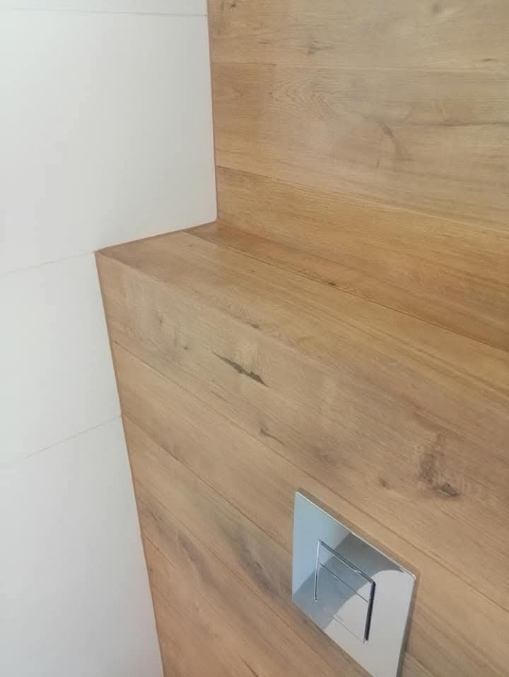 Fragment łazienki: drewnopodobna okładzina ścienna z przyciskiem spłuczki, białe płytki. Minimalistyczny design, ciepłe odcienie drewna, nowoczesne wykończenie.