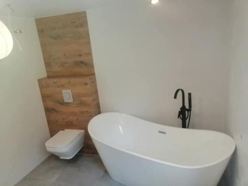 Nowoczesna łazienka z wolnostojącą białą wanną i toaletą w zabudowie z drewnopodobnych paneli, czarna armatura, szare płytki podłogowe, minimalistyczny design.