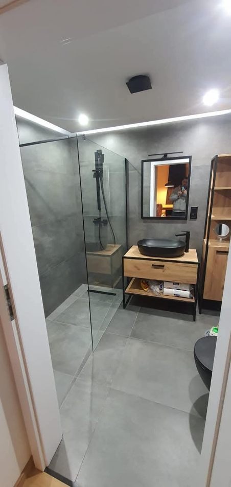 Nowoczesna łazienka z kabiną typu walk-in, szarą armaturą i drewnianą szafką pod umywalką. W lustrze odbija się osoba robiąca zdjęcie.