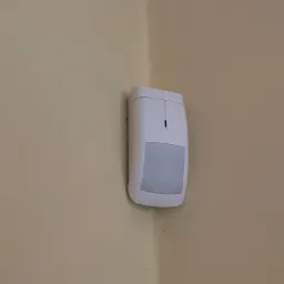 Biały czujnik ruchu alarmowego zamontowany w rogu beżowej ściany.