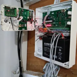 Otwarta obudowa centrali alarmowej z akumulatorem i okablowaniem, zamontowana na drewnianej ścianie w Konotopie, widoczne diody sygnalizacyjne i elementy elektroniczne.