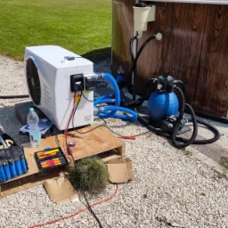 Instalacja pompy ciepła do basenu ogrodowego, widoczne niebieskie węże, filtr piaskowy, narzędzia w skrzynce oraz butelka wody obok urządzenia.