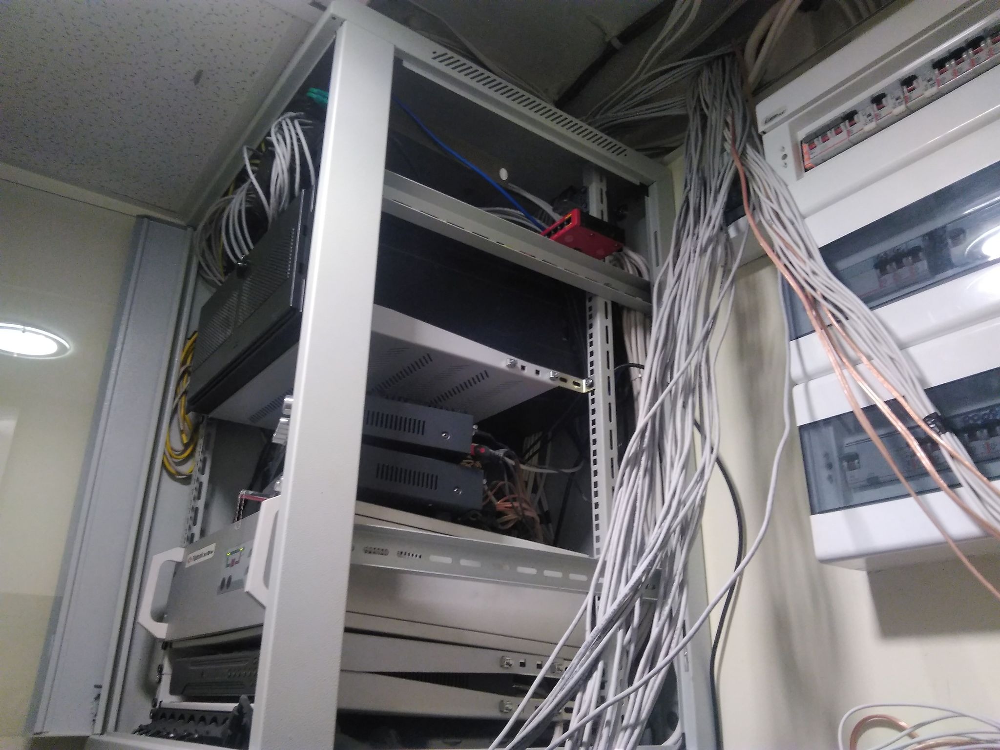 Szafa rack z urządzeniami sieciowymi i plątaniną kabli, widoczna skrzynka z bezpiecznikami po prawej stronie.