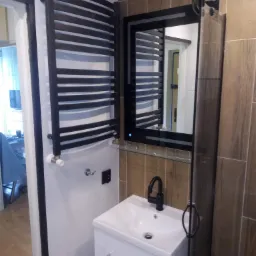 Nowoczesna łazienka z białą umywalką na szafce, czarną baterią, grzejnikiem drabinkowym i lustrem z podświetleniem LED, wykończona płytkami imitującymi drewno i białymi płytkami.