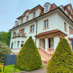 Elewacja dwupiętrowego budynku mieszkalnego z czerwonym dachem i pastelowymi detalami architektonicznymi, widziana z poziomu chodnika, z dwoma stożkowymi krzewami na pierwszym planie.