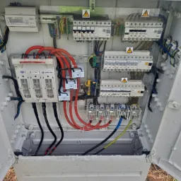 Otwarta skrzynka elektryczna z bezpiecznikami, przewodami i licznikiem energii, widoczne oznaczenia ostrzegawcze o napięciu.