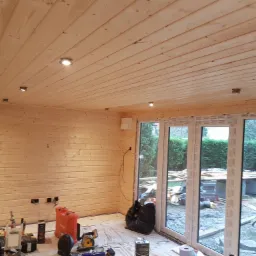 Wnętrze drewnianego domku z widocznymi deskami na ścianach i suficie, małymi lampkami w suficie, dużymi oknami z widokiem na ogród oraz narzędziami i materiałami budowlanymi na podłodze...