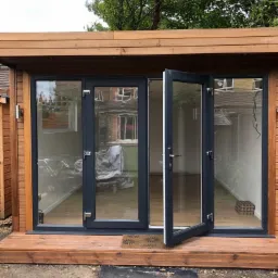 Nowoczesna drewniana altana z dużymi, grafitowymi oknami i częściowo otwartymi drzwiami, umieszczona na drewnianym podeście z brązową wycieraczką. Widoczne odbicie sąsiedniego budynku w szybach.