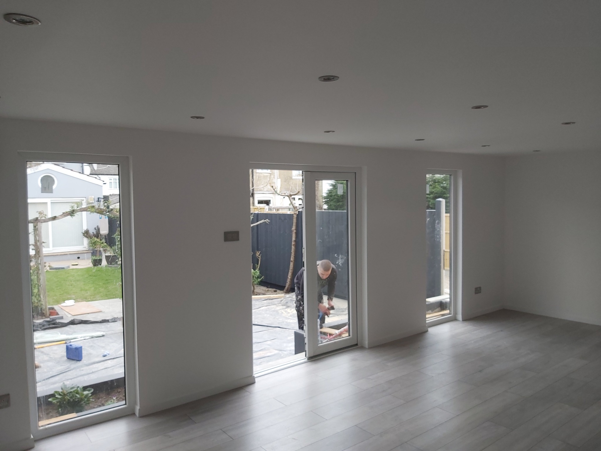 Wnętrze świeżo wyremontowanego pokoju z szarą podłogą, białymi ścianami i trzema oknami. Przez środkowe okno widoczna osoba pracująca na zewnątrz.