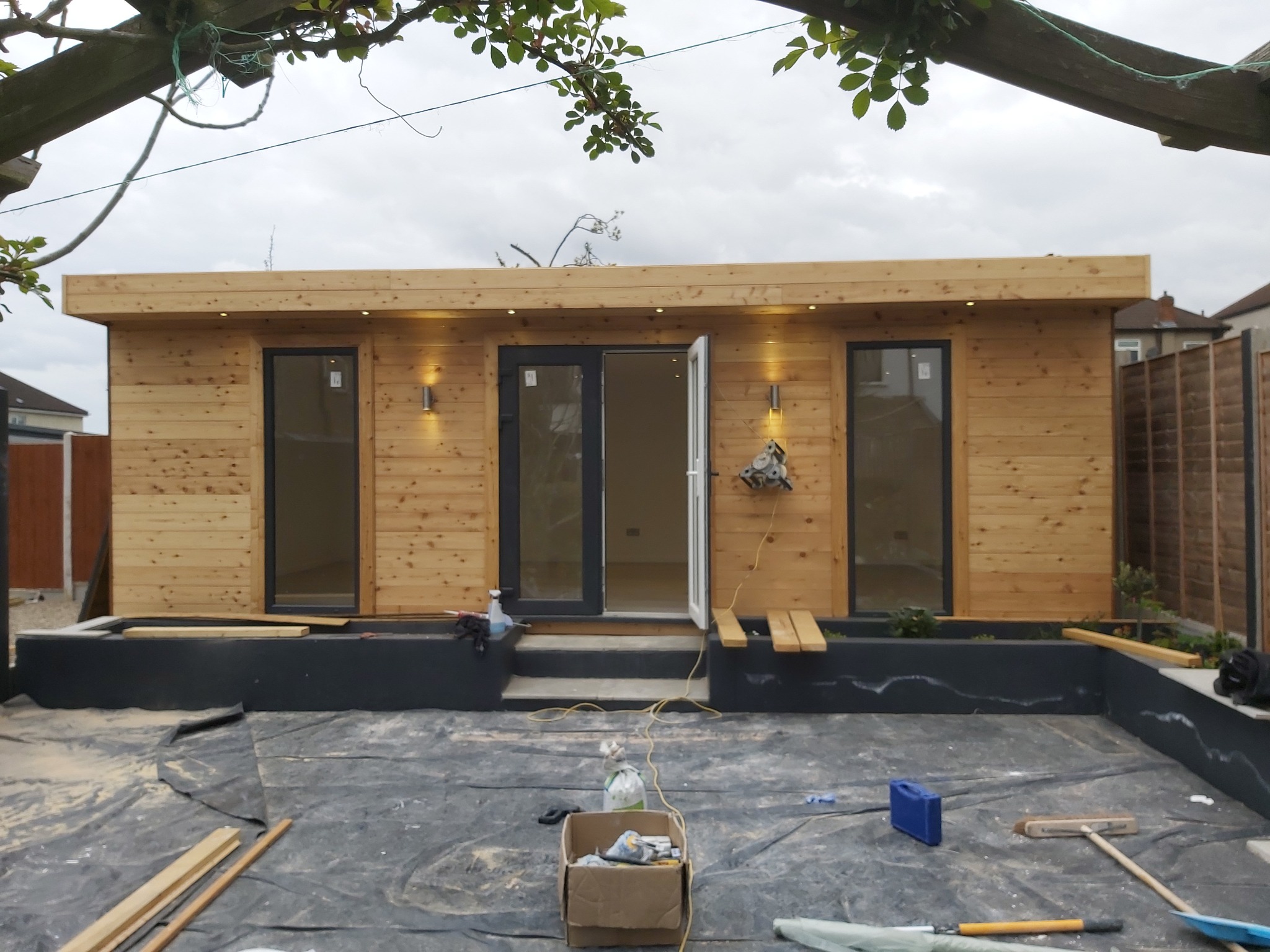 Nowoczesny drewniany domek ogrodowy z oświetleniem zewnętrznym w trakcie budowy, widoczne narzędzia i folia ochronna na ziemi.