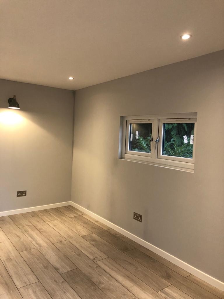Wnętrze pokoju po remoncie: świeżo pomalowane szare ściany, jasna drewnopodobna podłoga, białe listwy przypodłogowe, okno z widokiem na zieleń, dwa gniazdka elektryczne i lampa ścienna.