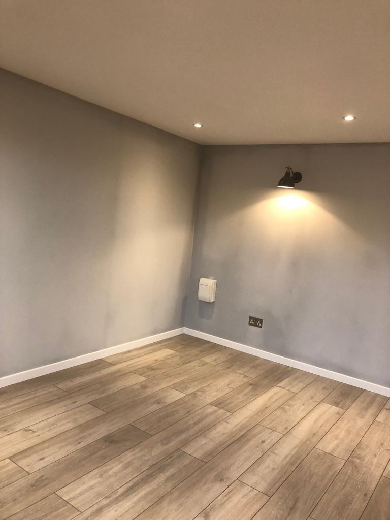 Pomalowane na szaro wnętrze pokoju z drewnopodobną podłogą, listwami przypodłogowymi, kinkietem i gniazdkami elektrycznymi.
