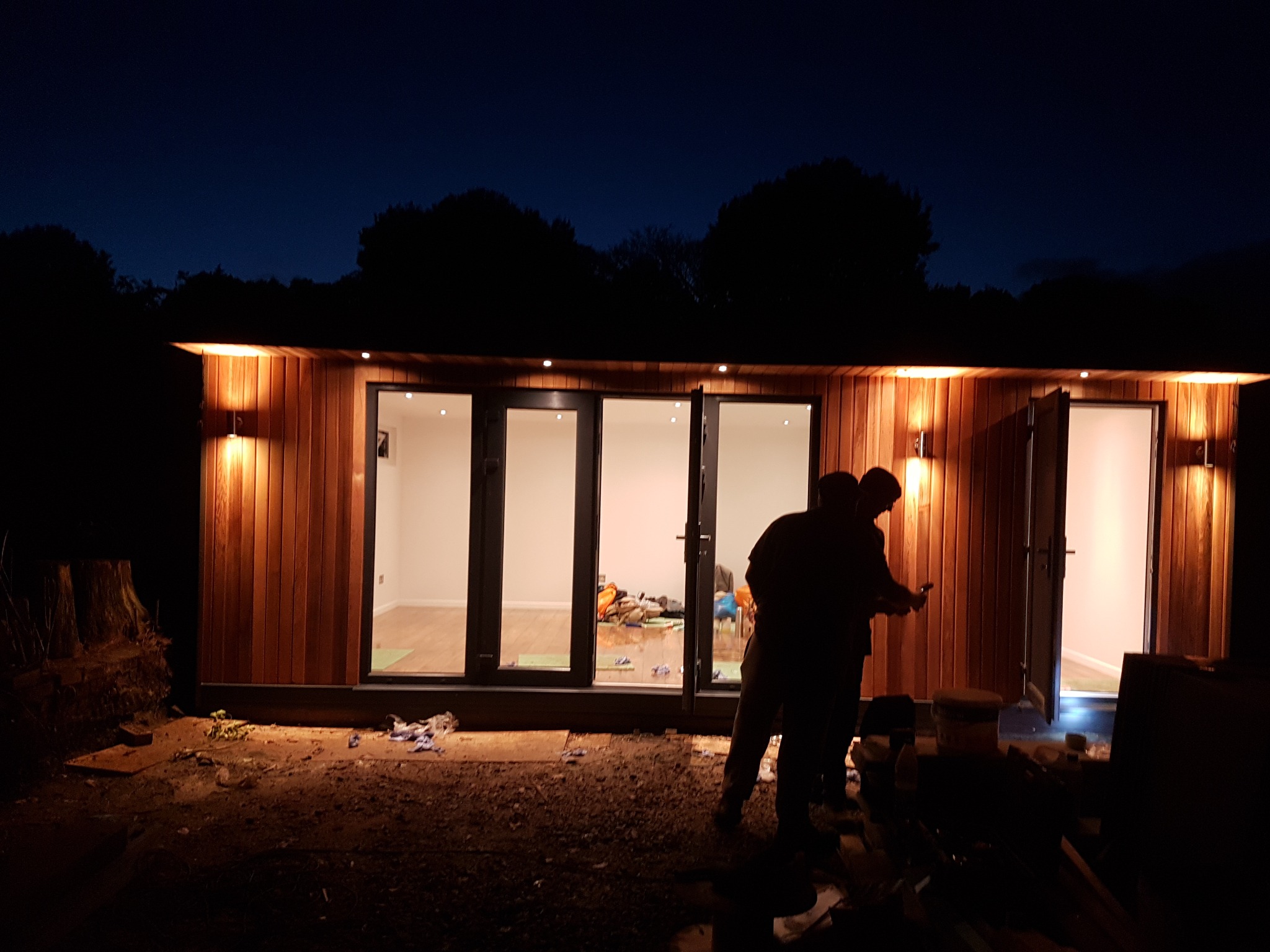 Oświetlony, drewniany budynek gospodarczy z dużymi oknami i otwartymi drzwiami wejściowymi, widziany w nocy, z dwoma osobami stojącymi przed nim, prawdopodobnie podczas prac wykończeniowych.