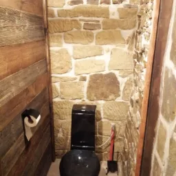 Mała łazienka z czarną toaletą, ściany wykończone kamieniem i drewnem, widoczny podajnik na papier toaletowy i szczotka do toalety.