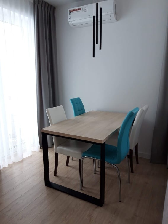 Jasne wnętrze jadalni z dębowym stołem na czarnej, metalowej podstawie, otoczone krzesłami w kolorze białym i turkusowym, oświetlone wiszącymi, czarnymi lampami tubowymi i klimatyzatorem.