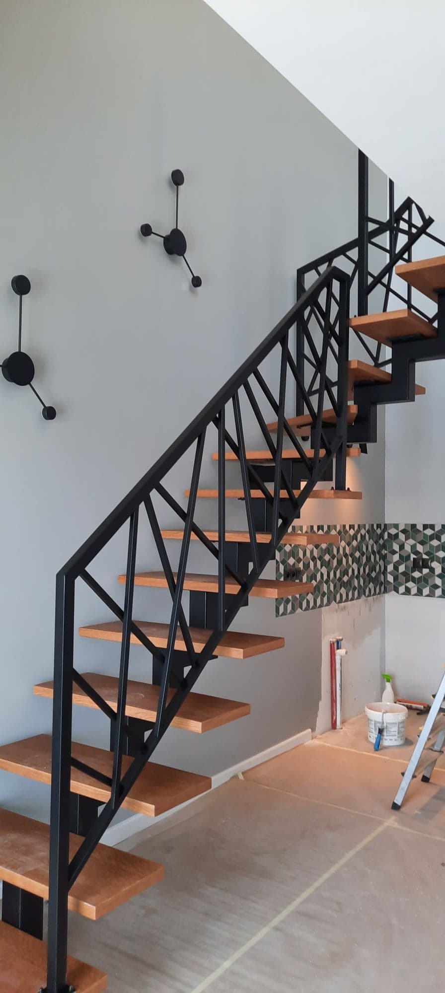 Nowoczesne schody z drewnianymi stopniami i czarną, geometryczną balustradą, na tle szarej ściany z trzema czarnymi zegarami w stylu molekularnym, widoczne prace wykończeniowe przy ścianie z mozaiką.