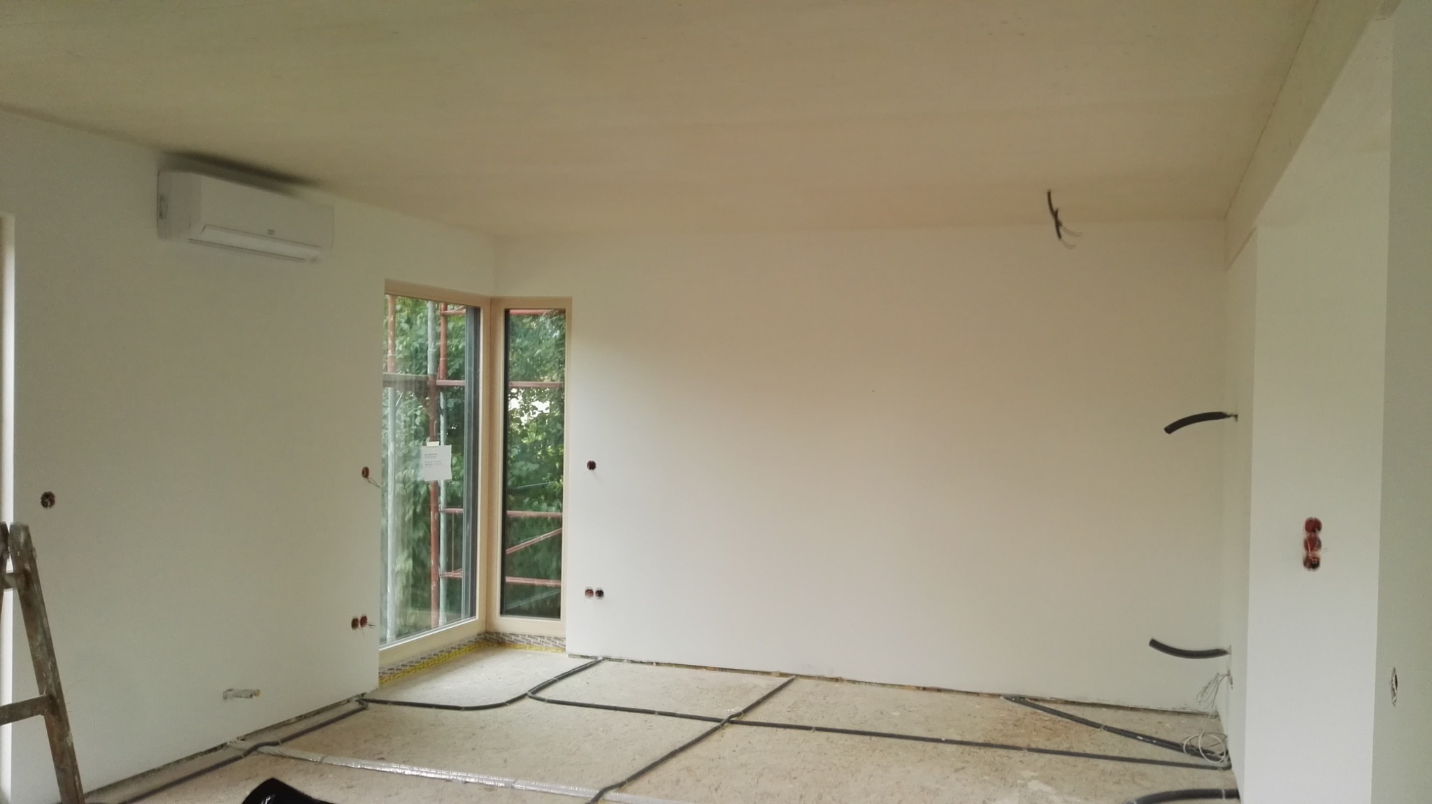 Wnętrze w trakcie prac wykończeniowych: białe ściany, okno narożne z widokiem na zieleń, klimatyzator na ścianie, instalacja elektryczna i rury grzewcze ułożone na podłodze, drabina oparta o ścianę.