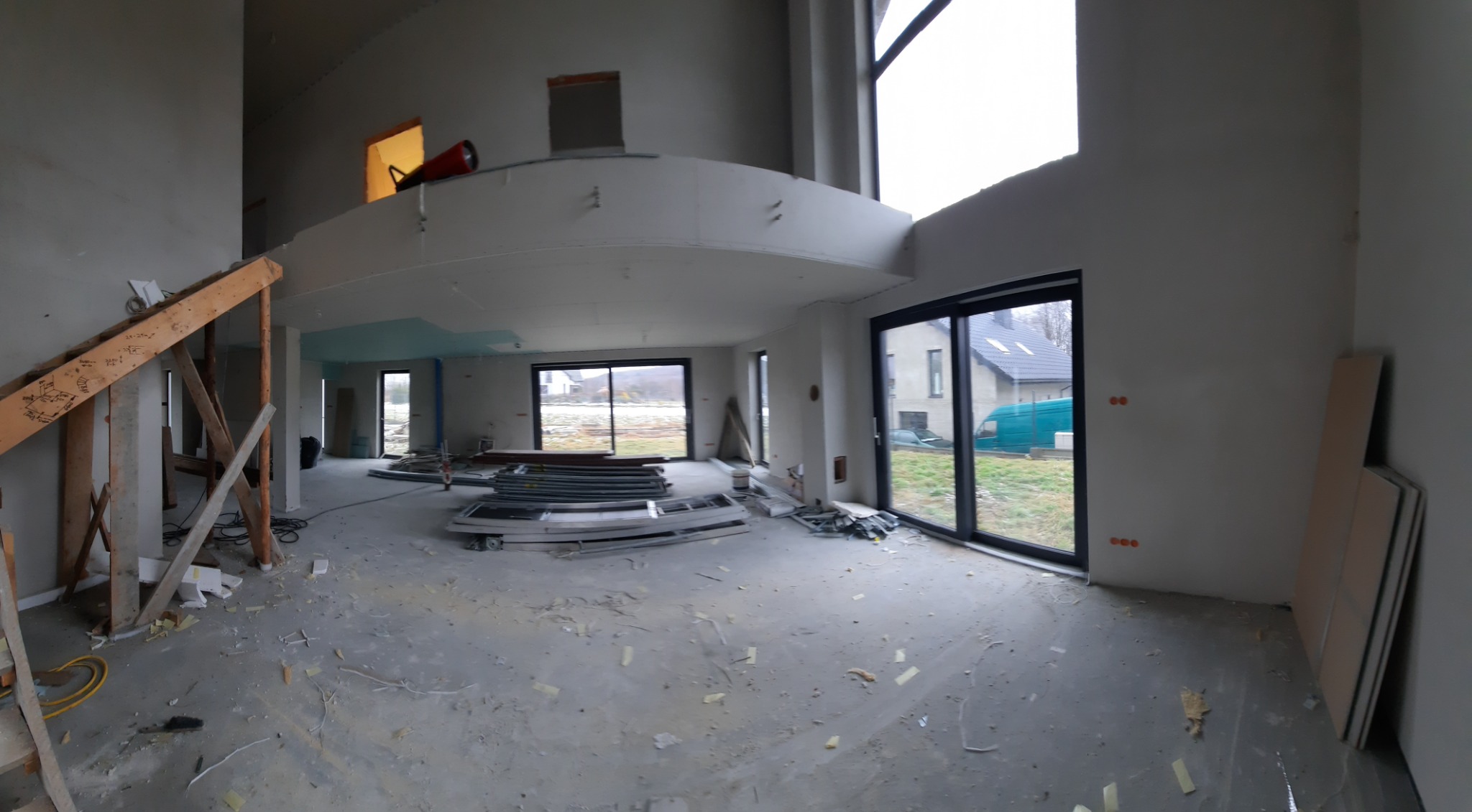 Wnętrze domu w trakcie remontu, widoczne surowe ściany, podłoga pokryta resztkami materiałów budowlanych, przygotowane okna i elementy drewniane konstrukcji schodów.