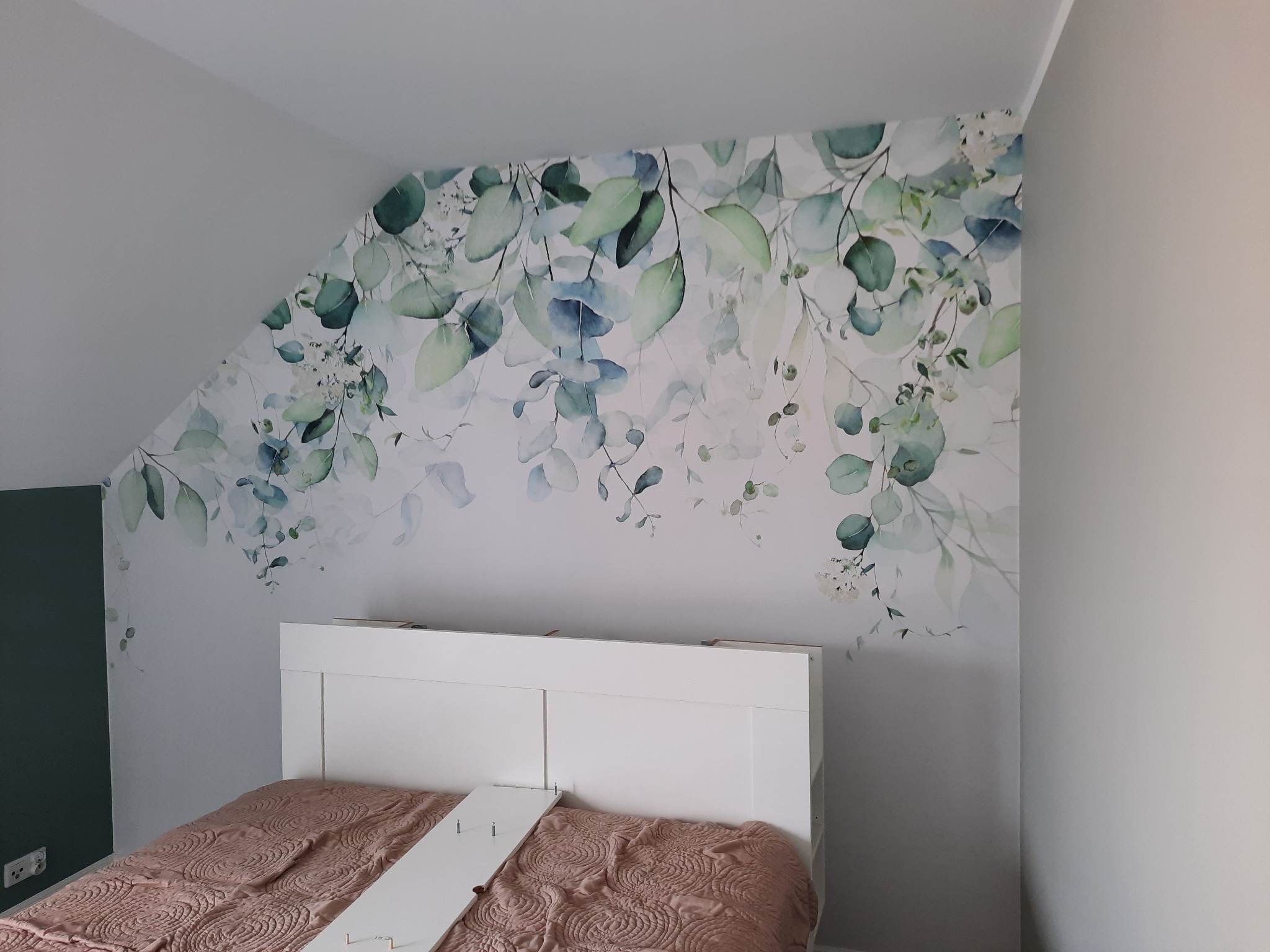 Ściana w pokoju na poddaszu z fototapetą w motywy roślinne, białe wezgłowie łóżka i fragment zielonej ściany z gniazdkiem elektrycznym.