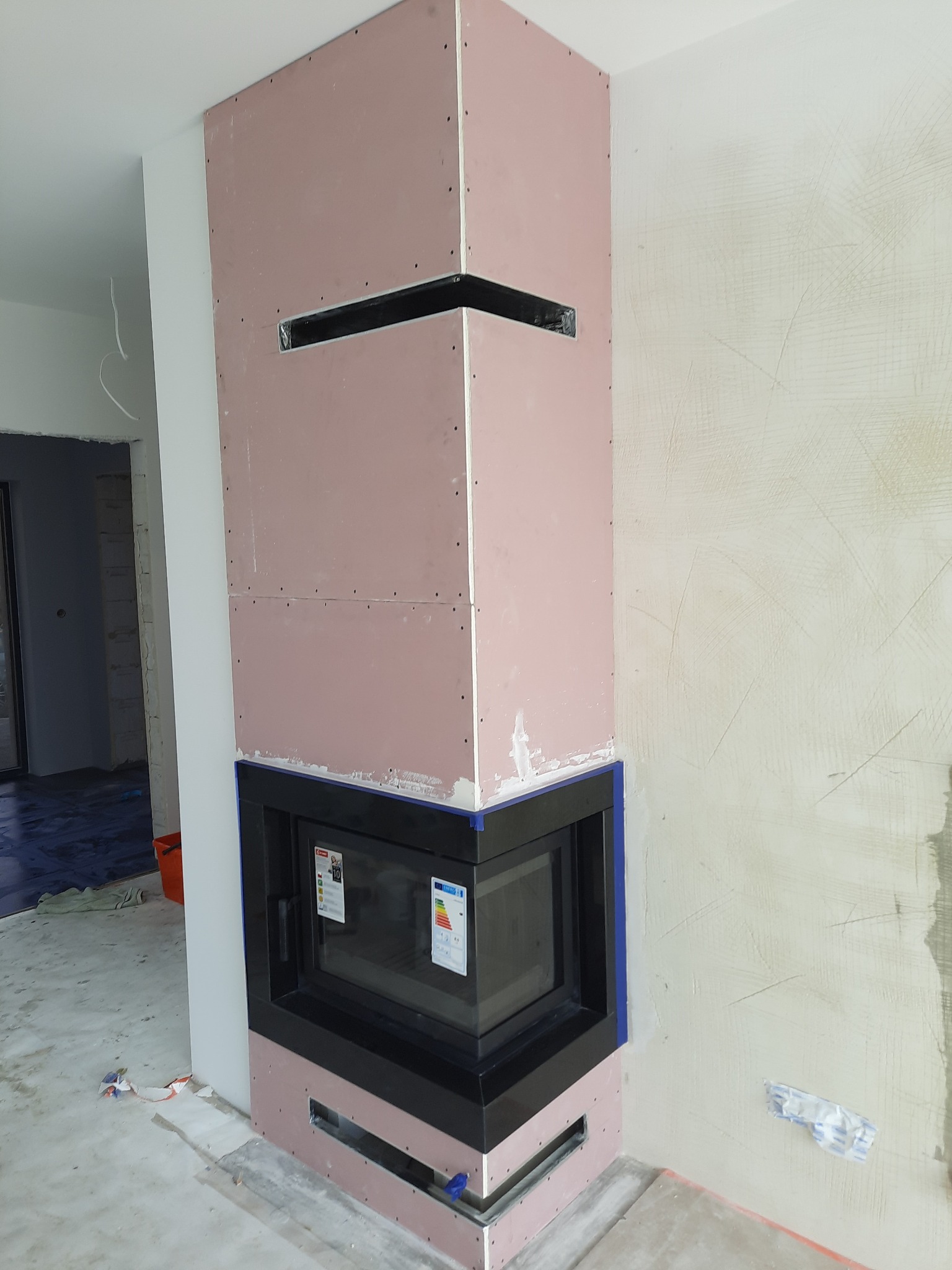 Wnęka kominkowa w trakcie obudowy płytami gipsowo-kartonowymi, widoczne otwory wentylacyjne i czarna rama wkładu kominkowego, ściany w stanie surowym przed malowaniem.